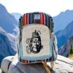 Handmade cotton and hemp backpack with Lord Shiva print, featuring colorful Himachali tribal patterns, placed on a rock with a scenic Himalayan lake and mountains in the background